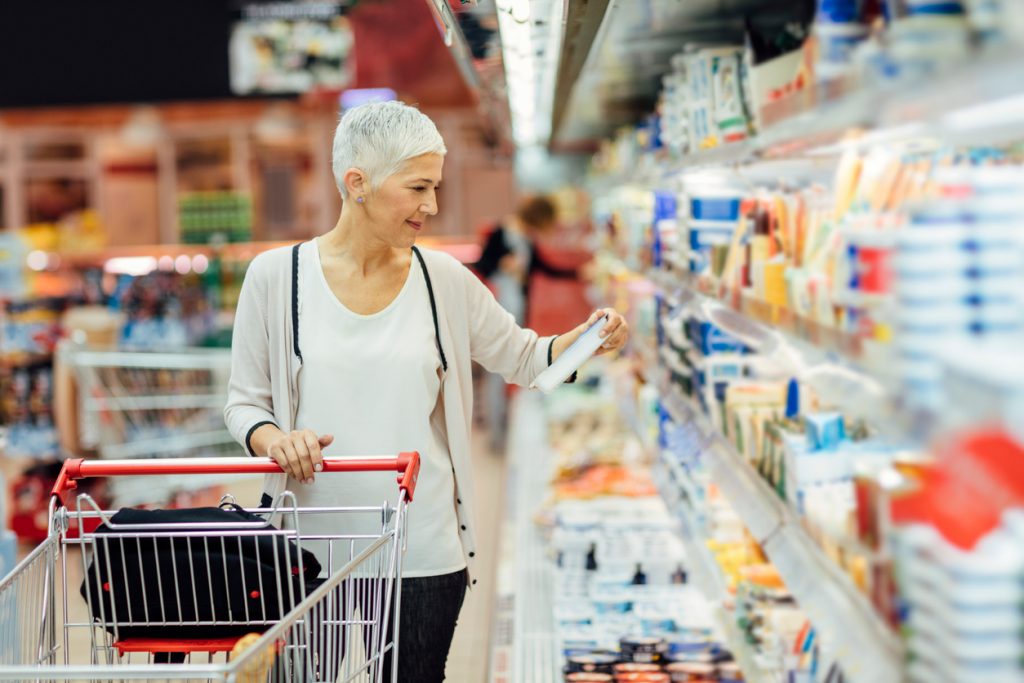 Mature smiling woman shopping in local supermarket. She is shopping groceries. Standing by produce stand and reading nutrition label on dairy product. Choosing cheese.
